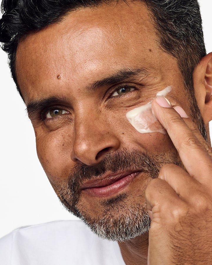 Close up of smiling, bearded man applying Clinique For Men Anti-Age Eye Cream to under eyes, in a bright setting